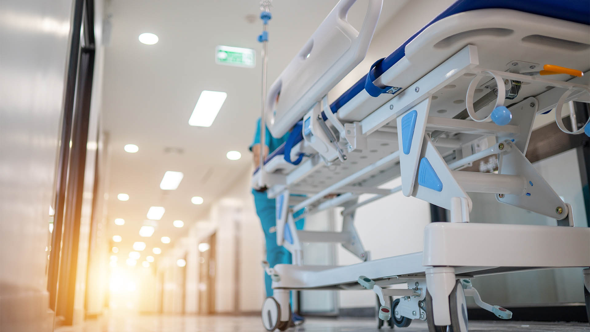 A hospital room with a medical staff member pushing a hospital bed. The room is bright and sterile, with a large window letting in natural light. The medical staff member is wearing a blue uniform