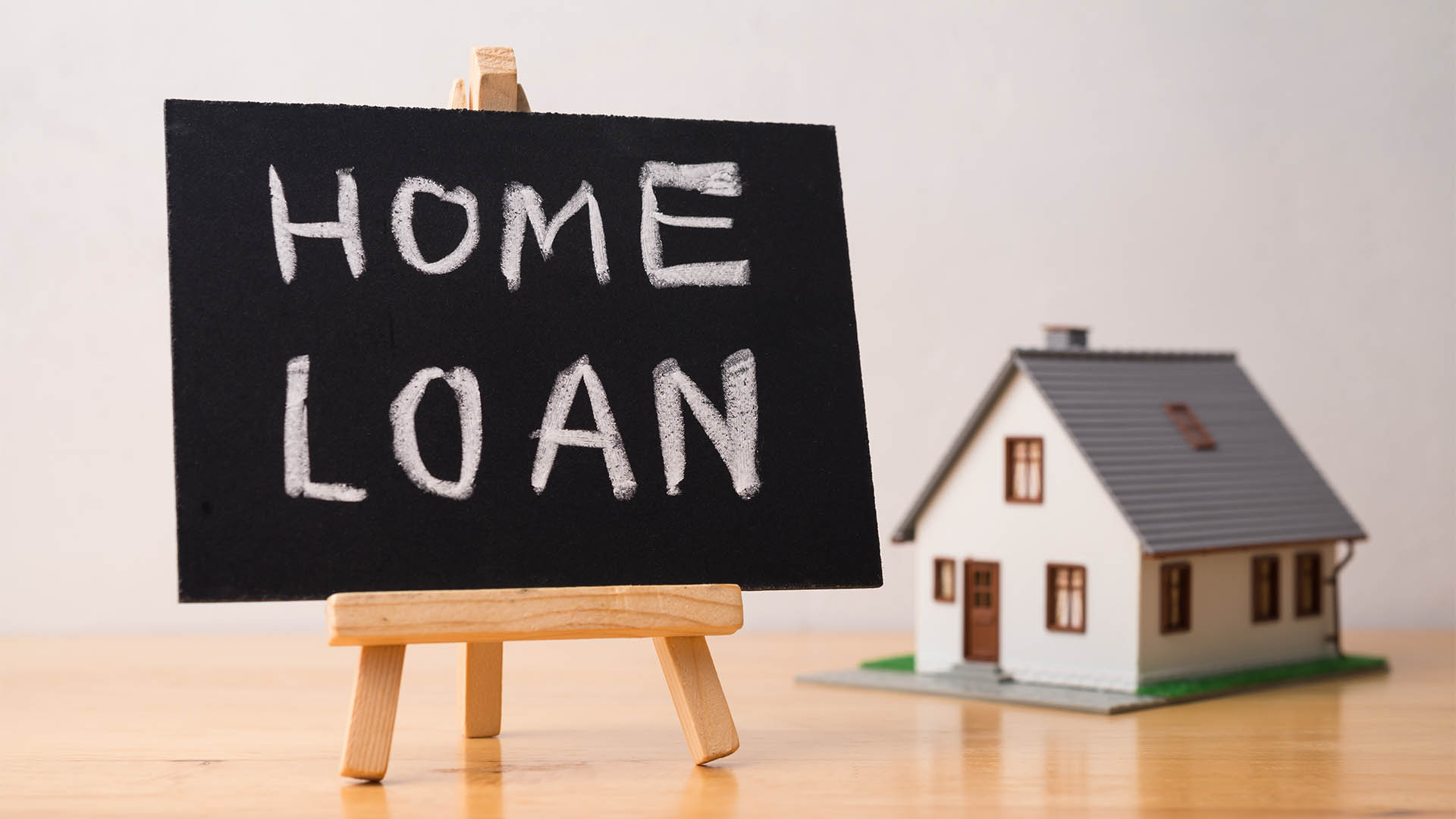 Home loan is written on small blackboard and house model on wooden table with white wall background. Home loan, mortgage loan, personal financial, buying home, banking and refinancing concept.