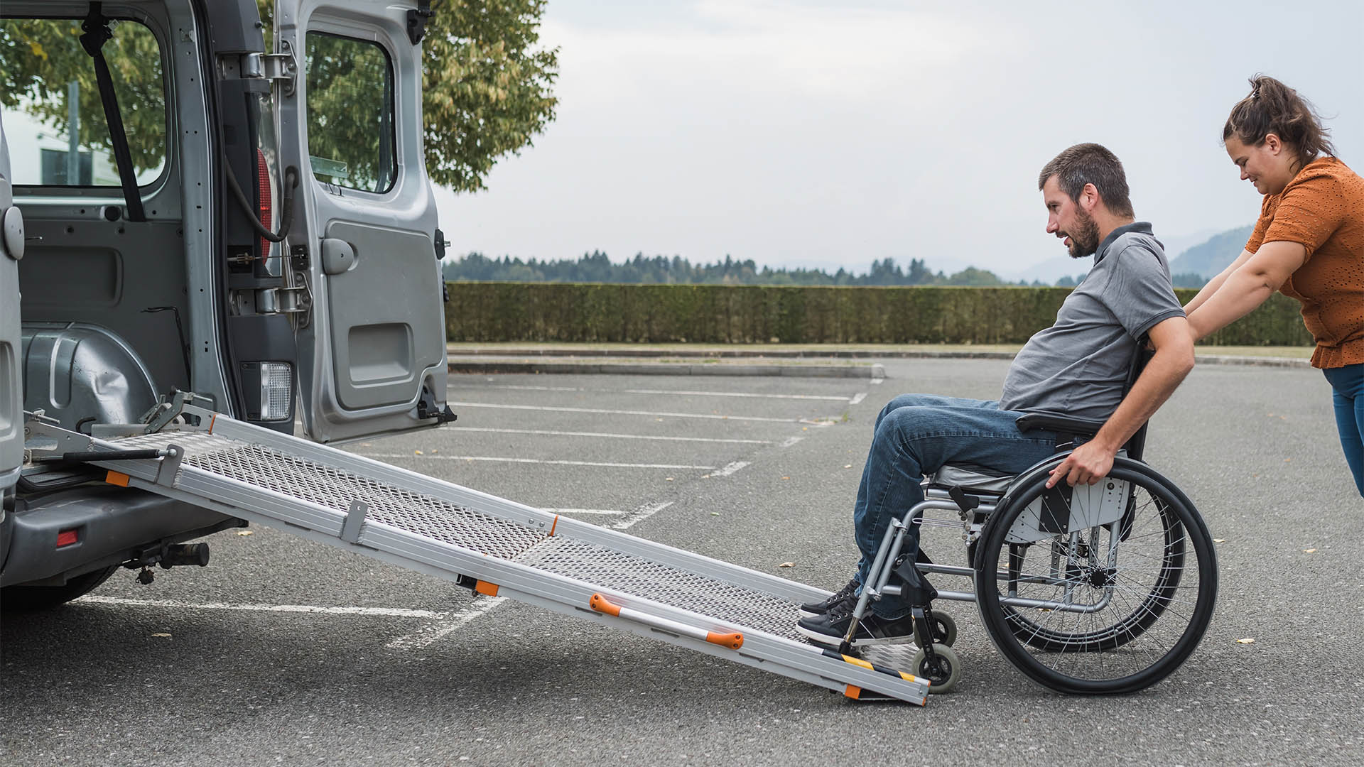 Female assistant pushing a man with disability, a wheelchair user up the car ramp into the van. Accessibility and travel concepts.