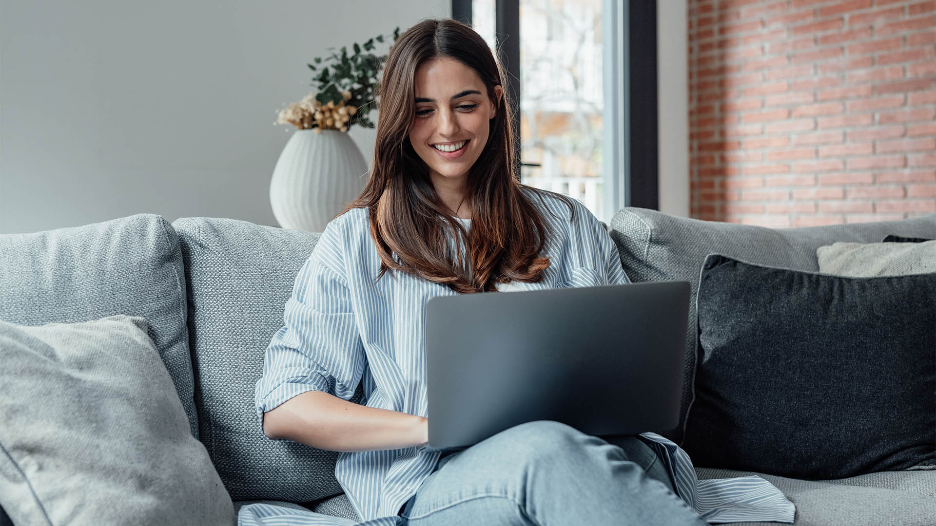 Concentrated millennial girl sit on couch working on laptop browsing internet at home during lazy weekend, focused young woman freelancer busy using computer surfing wireless web shopping online