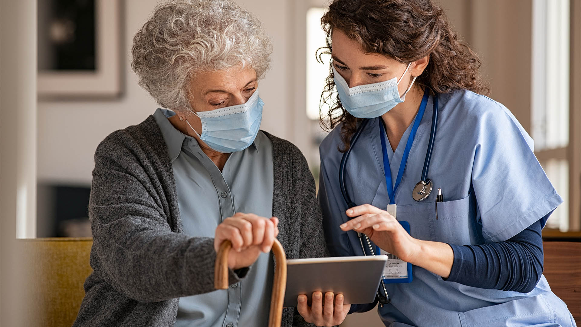 Doctor and senior woman going through medical record on digital tablet during home visit wearing face mask. Old woman with nurse with surgical mask and using digital tablet during coronavirus pandemic