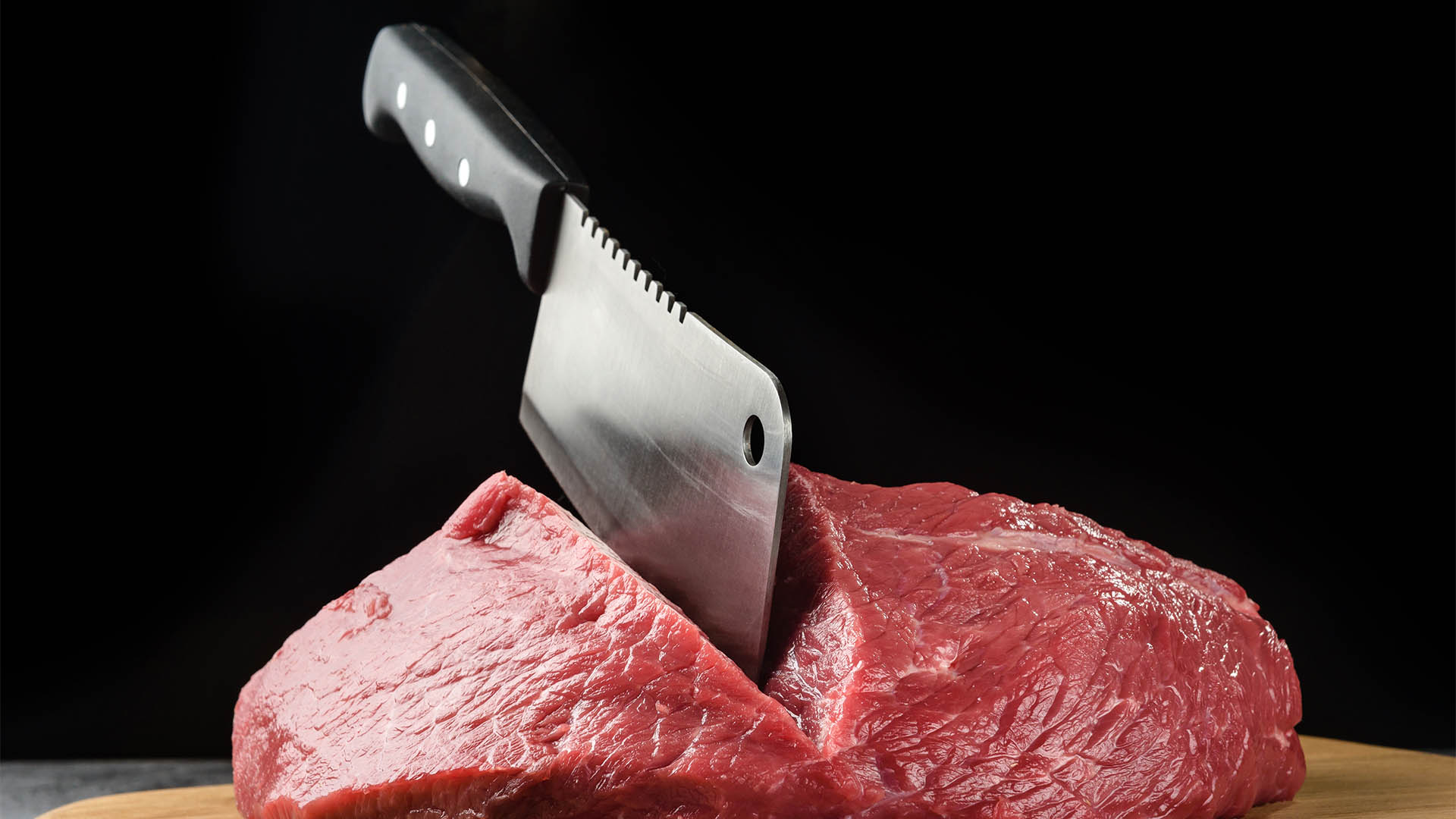 A piece of raw meat close-up with a chef's hatchet on a dark background.