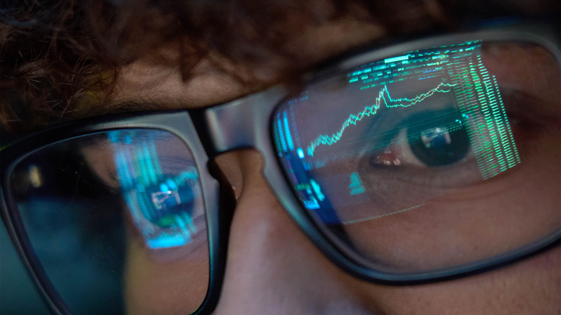 Young indian business man trader wearing glasses looking at computer screen with trading charts reflecting in eyeglasses watching stock trading market financial data growth concept, close up.